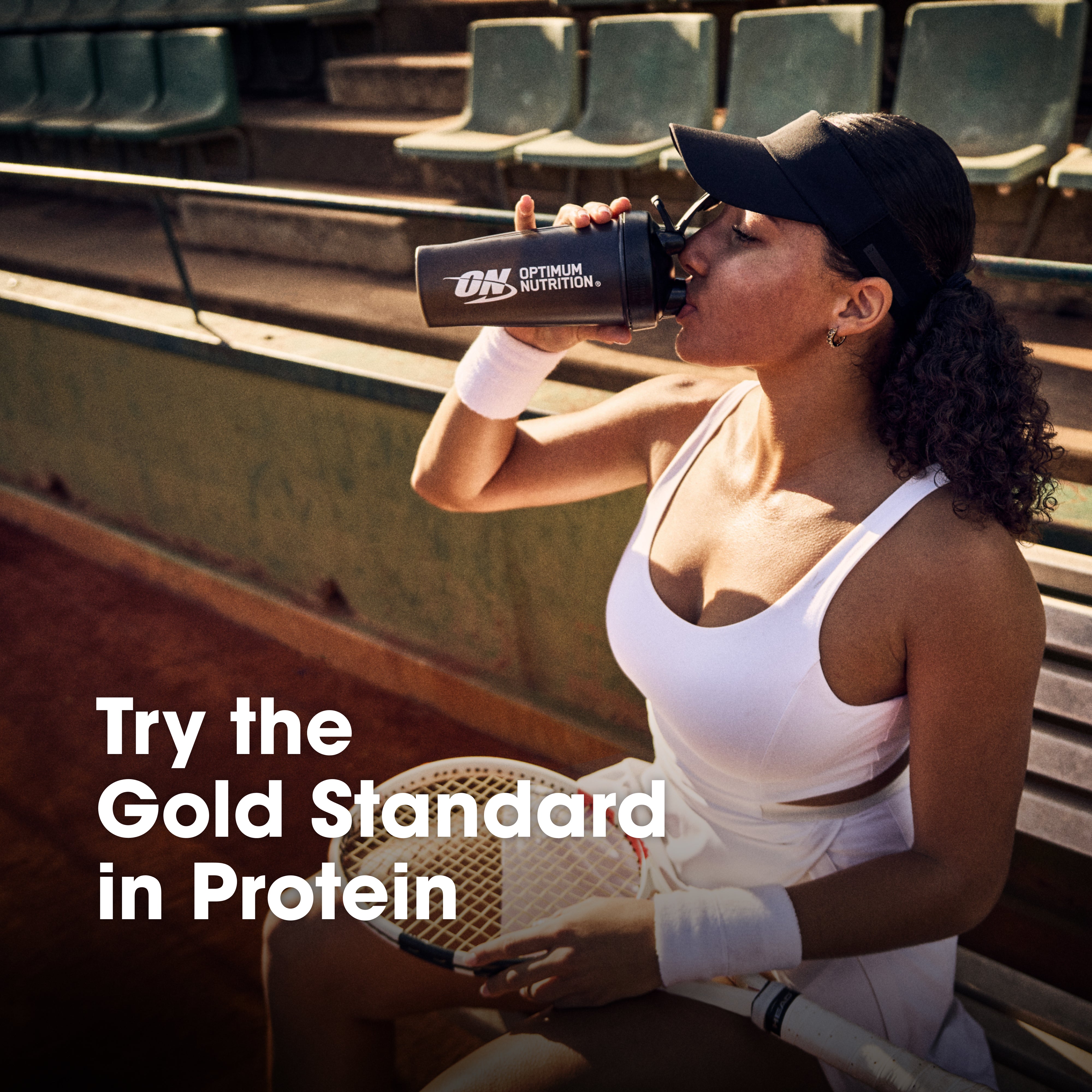 A woman in a white tennis outfit sits on stadium seating holding a protein shaker bottle with the Optimum Nutrition logo. Large text on the image reads 'Try the Gold Standard in Protein'.
