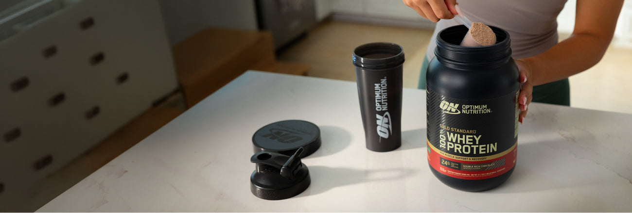 Person preparing a protein shake with Optimum Nutrition whey protein powder on a kitchen counter.